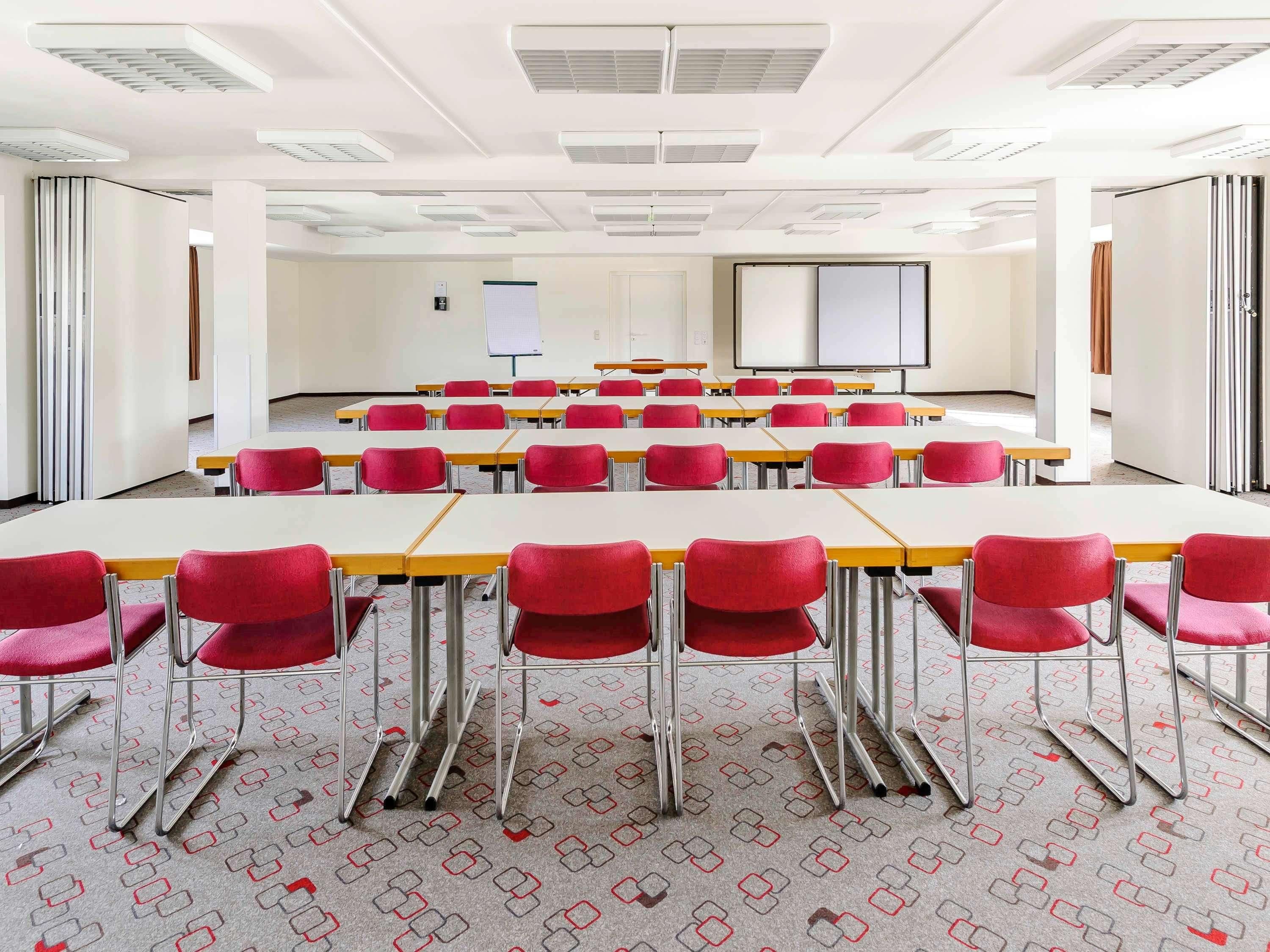 A conference room with rows of tables and chairs, white walls, and a projector screen.
