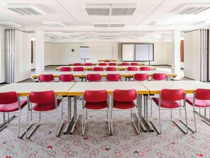 Un salón de conferencias con filas de mesas y sillas, paredes blancas y una pantalla de proyector.