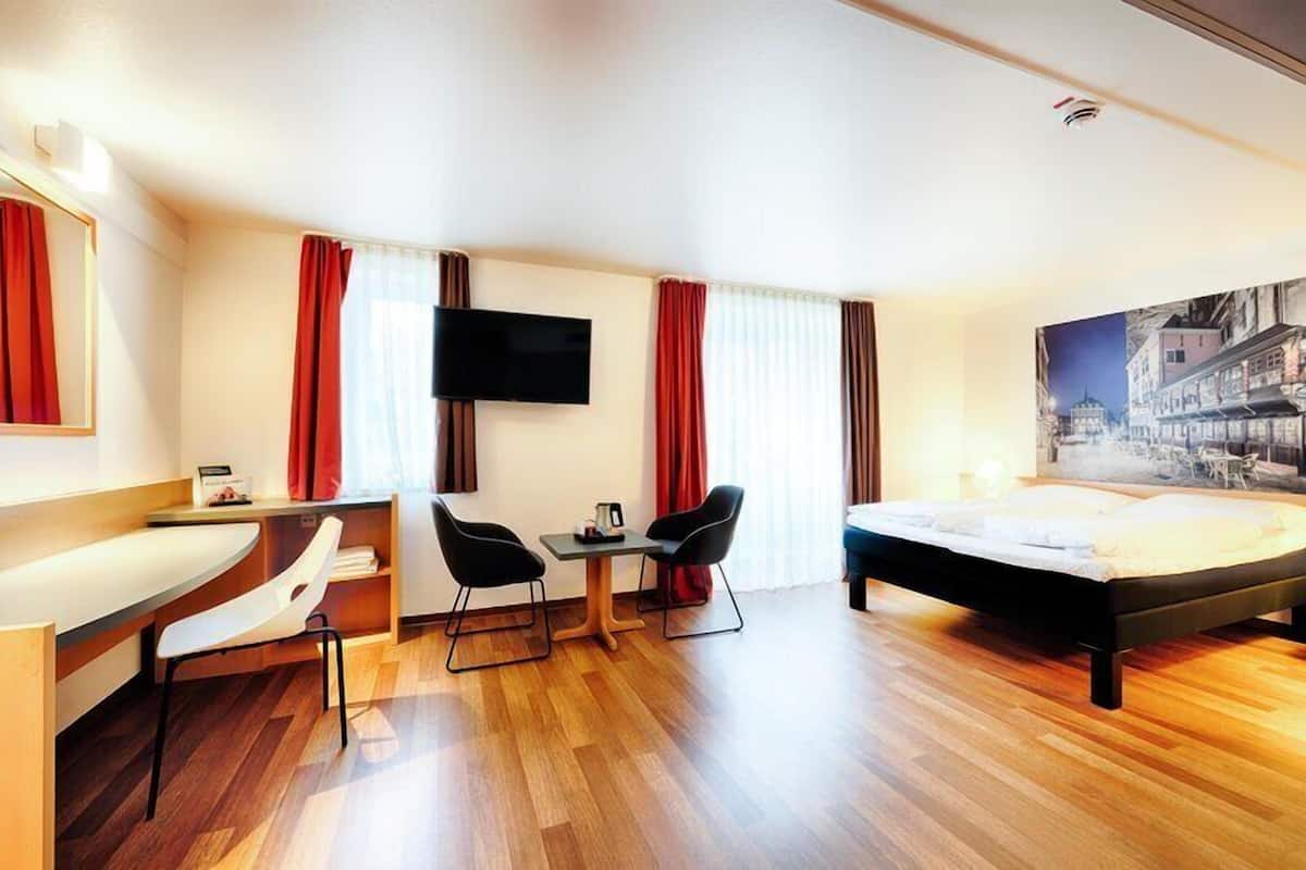 A hotel room with a bed, a desk with two chairs, a flat-screen TV, and a painting on the wall.