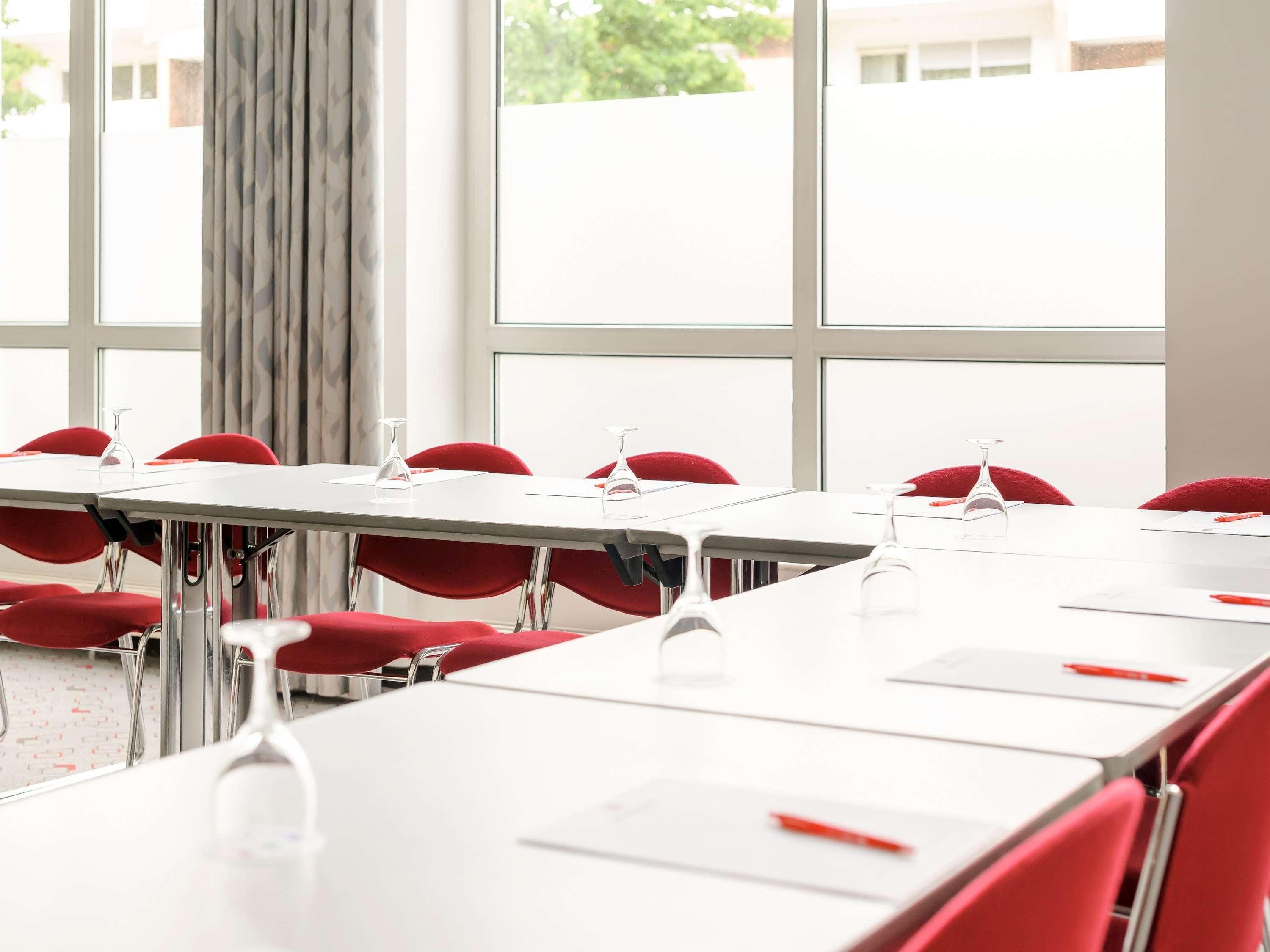 Un salón de conferencias con mesas blancas y sillas rojas, ventanas amplias y vista a edificios exteriores.