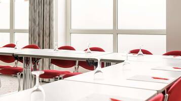 Un salón de conferencias con mesas blancas y sillas rojas, ventanas amplias y vista a edificios exteriores.