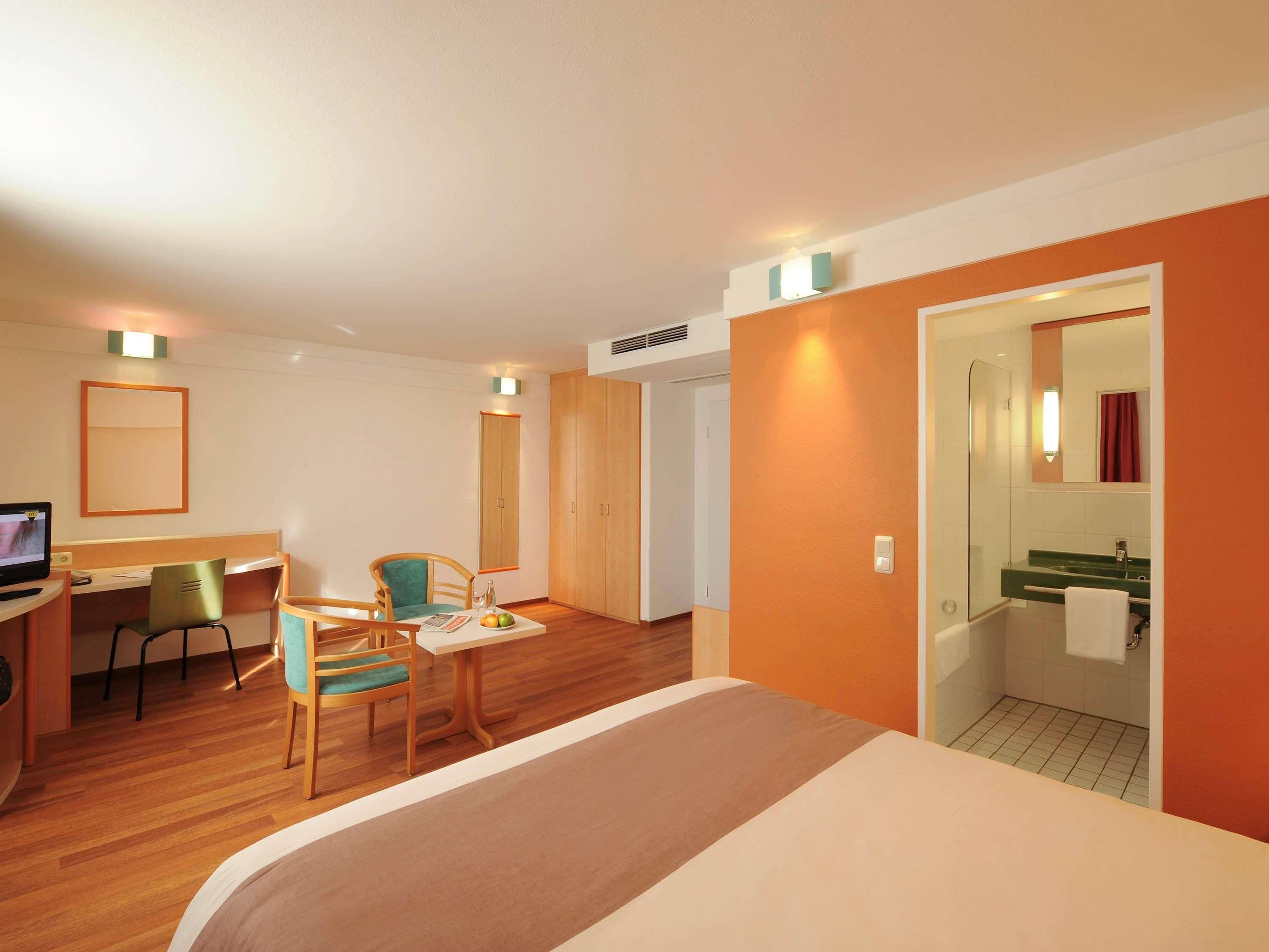 Habitación de hotel con una cama, un escritorio con computadora, dos sillas, una mesita con frutero, y baño con lavamanos y espejo.