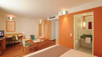 Habitación de hotel con una cama, un escritorio con computadora, dos sillas, una mesita con frutero, y baño con lavamanos y espejo.