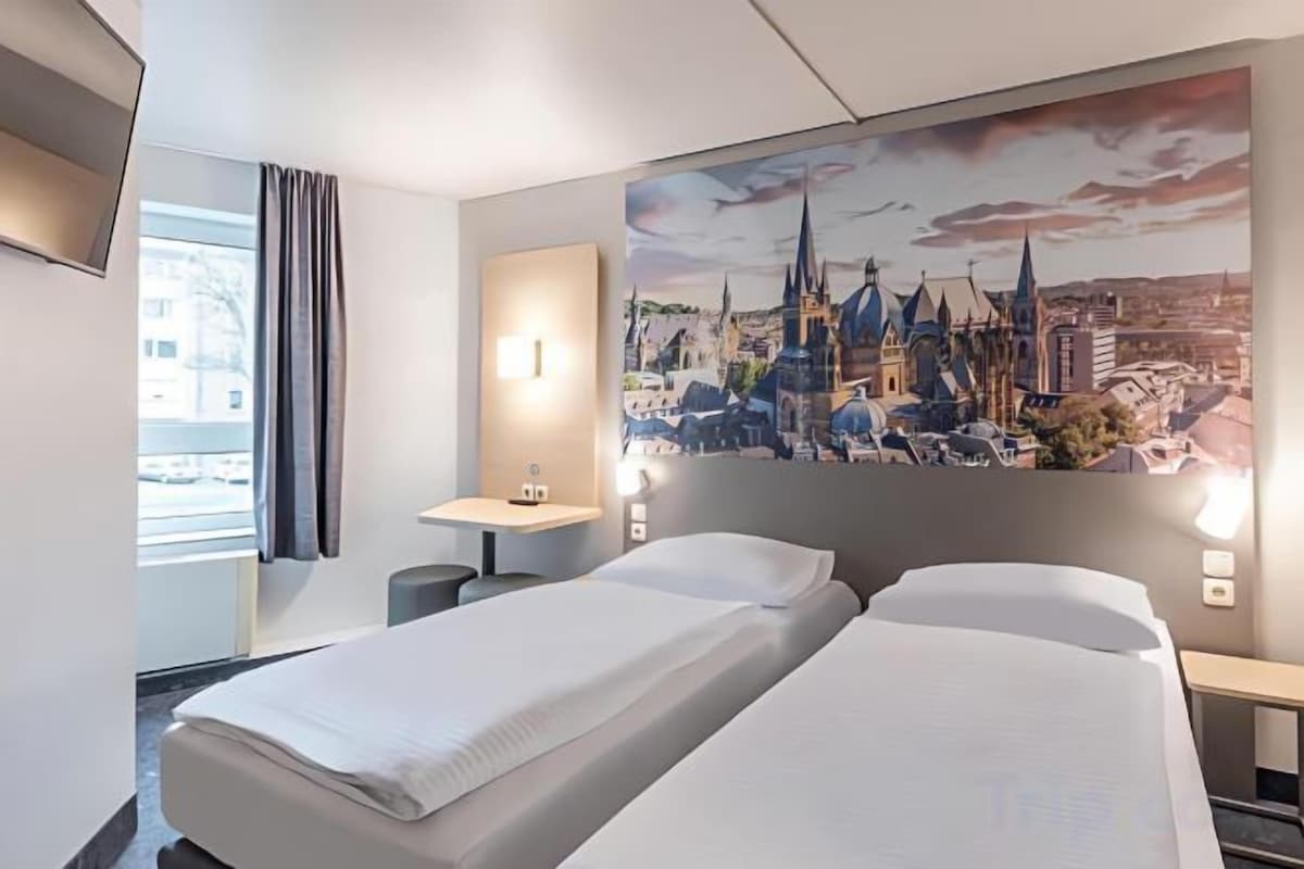 A hotel room with two beds, a nightstand, a lamp, and a cityscape painting on the wall.