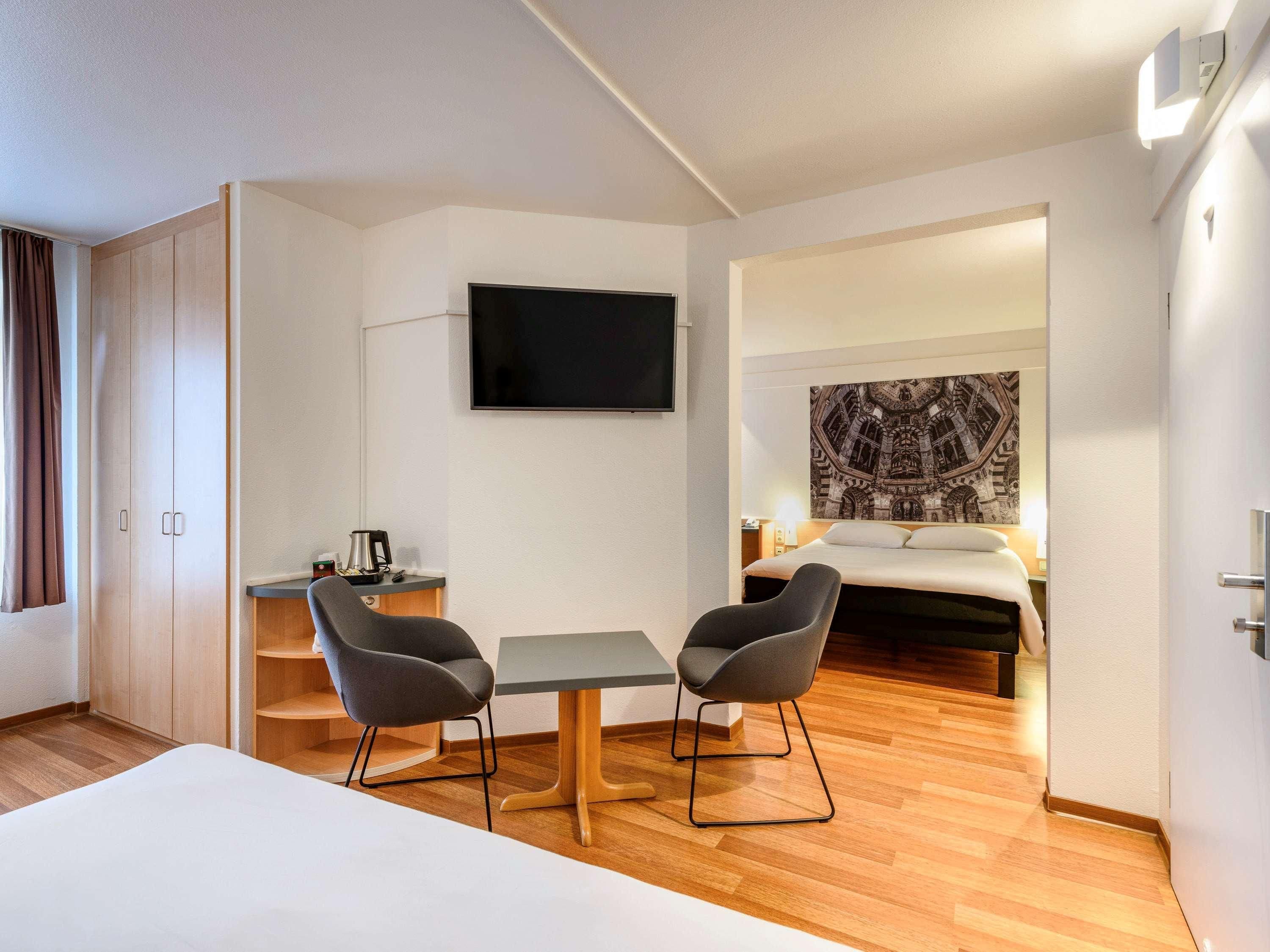 A hotel room with a bed, two chairs, a table, a TV, and a painting on the wall.