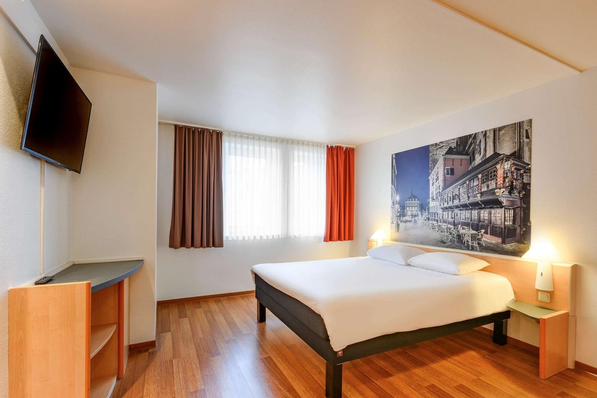 A hotel room with a bed, a desk, a TV, and a painting on the wall.