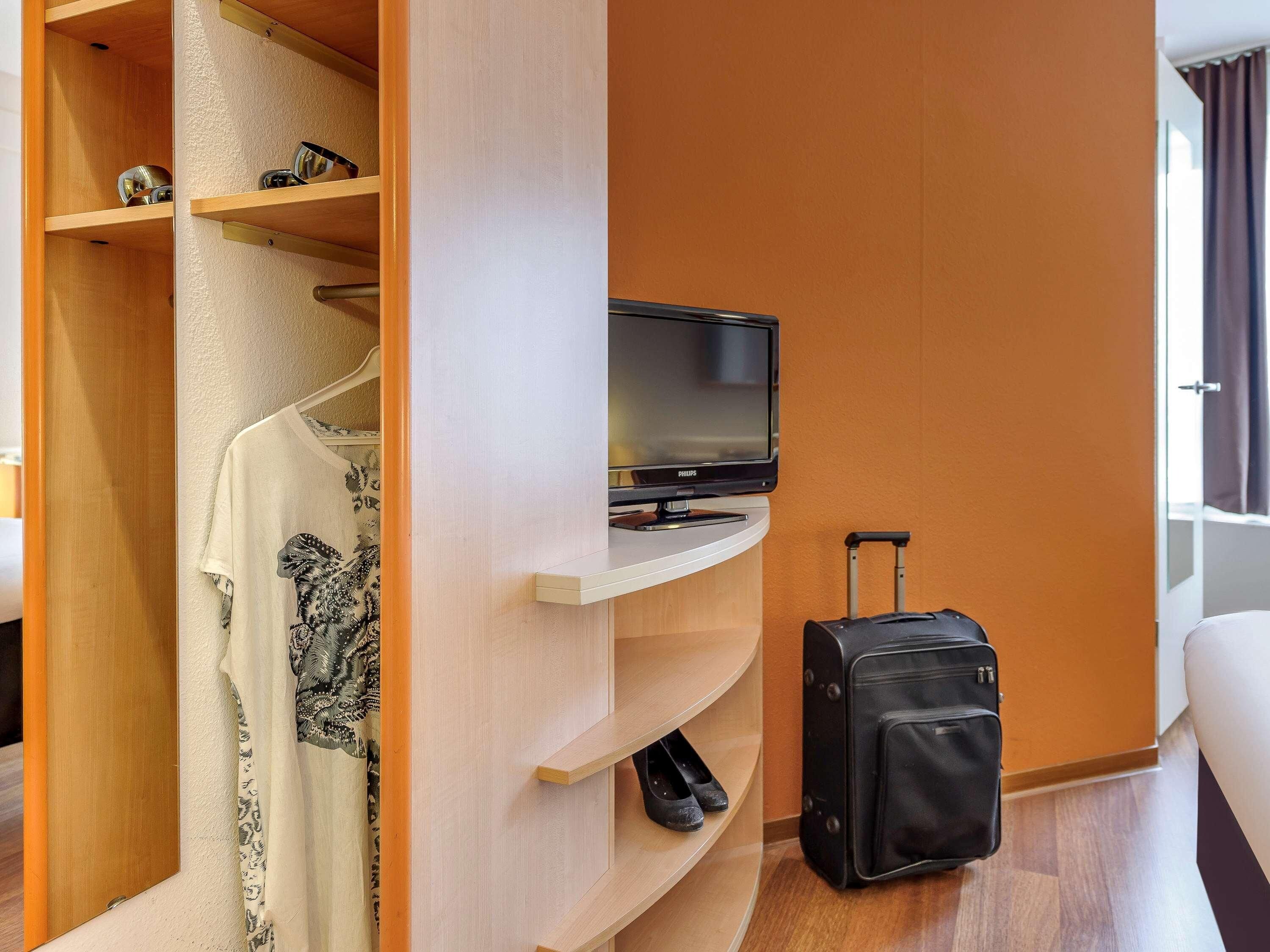 Habitación de hotel con televisor sobre un soporte, una maleta y un armario con estantes.