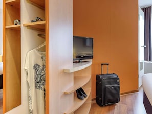 A hotel room with a television on a stand, a suitcase, and a wardrobe with shelves.