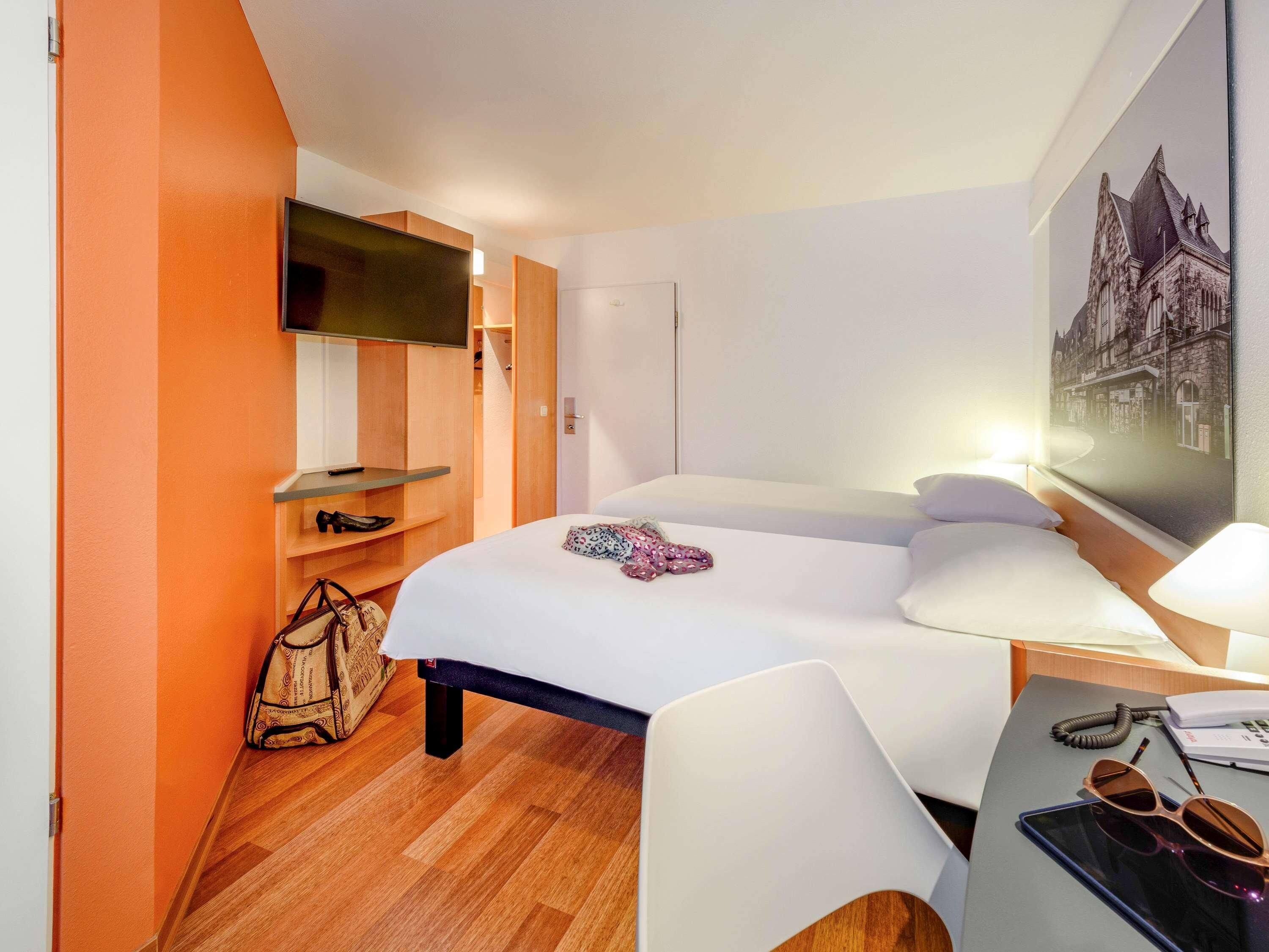 Habitación de hotel con cama, escritorio con lámpara, televisor montado en la pared y una pintura de paisaje urbano.