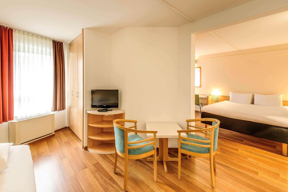 A hotel room with a bed, a desk with a TV, and two chairs.