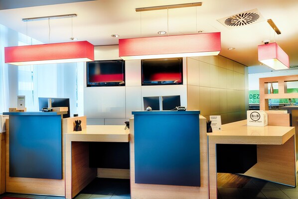 A modern reception desk area with three counters, each with a blue front panel and a wooden base. There are overhead red pendant lights and a ceiling vent. - B&B Aachen HBF (Aachen)