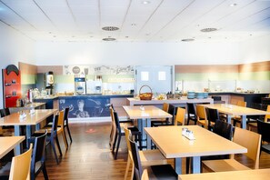 A cafe with wooden tables and chairs, a counter with a coffee machine, and a wall with a coffee-themed mural. - B&B Aachen HBF (Aachen)