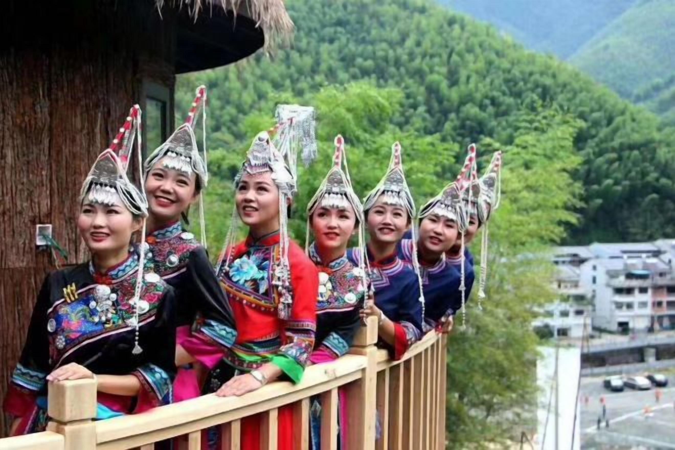 A group of people in traditional attire with elaborate headdresses, sitting on a wooden balcony with a mountainous landscape in the background.