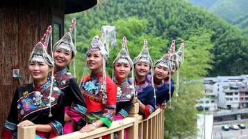 A group of people in traditional attire with elaborate headdresses, sitting on a wooden balcony with a mountainous landscape in the background.