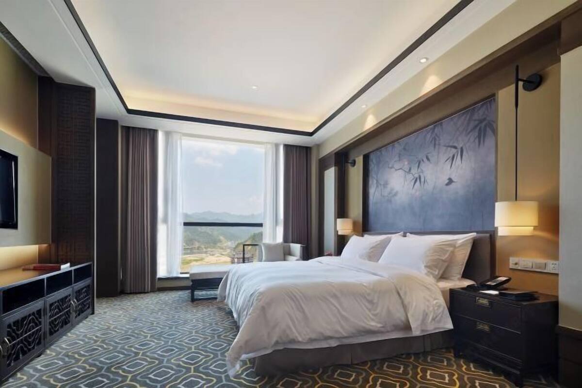 Habitación de hotel con cama, mesitas de noche, televisión y un ventanal con vistas al exterior.