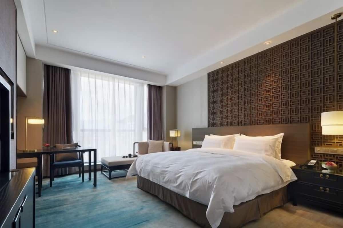 Habitación de hotel con una cama grande, un escritorio, una silla, un televisor y un papel tapiz estampado.