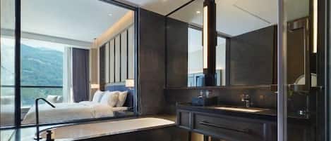 A modern hotel room with a large bed, a built-in bathtub, a vanity with a mirror, and a view of the mountains.