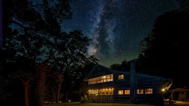 A two-story log cabin with a porch and multiple windows, surrounded by trees and a clear night sky.