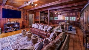 A rustic cabin interior with a large leather sofa, wooden beams, a fireplace, and a mounted television.