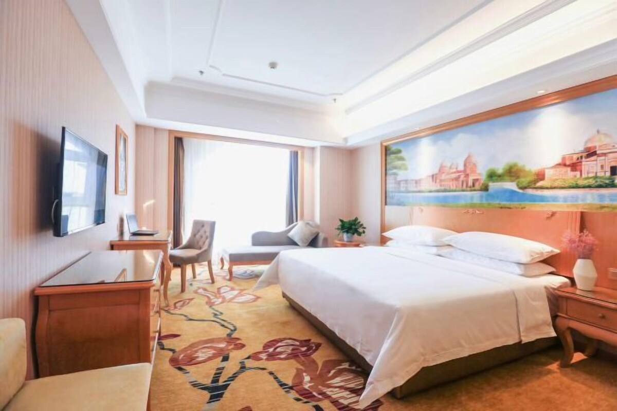 Ein Hotelzimmer mit Bett, Schreibtisch, Stuhl, Fernseher und einem Bild an der Wand.