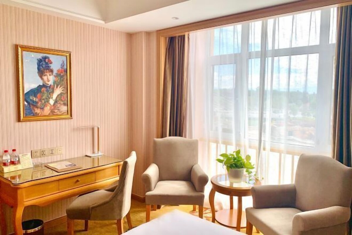 Ein Hotelzimmer mit Schreibtisch, zwei Sesseln, einem kleinen Tisch mit Pflanze, einem Bild an der Wand und großen Fenstern mit Vorhängen.