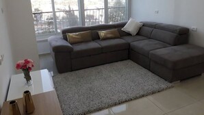 A modern living room with a sectional sofa, a small side table with a vase, and a large window with a view of buildings. - Heart of Jerusalem 4 rooms - modern - WIFI (Jerusalem)