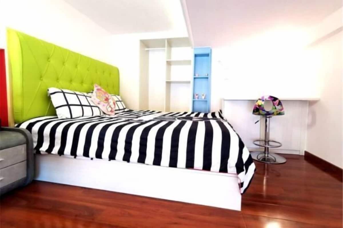 A bedroom with a tufted headboard, a striped bedspread, a grey nightstand, a blue shelving unit, and a small round table with a colorful umbrella.