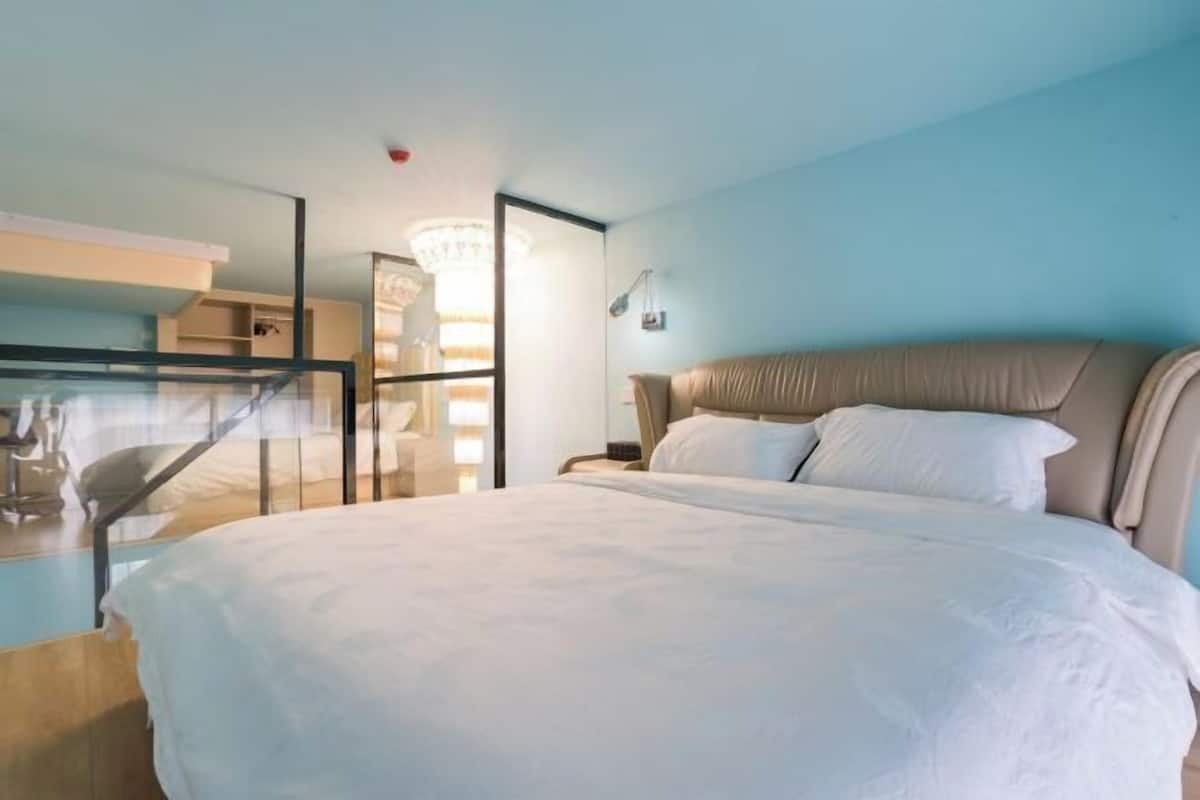 A hotel room with a large bed, a glass partition, a desk with a chair, and a chandelier.