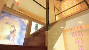 A staircase with wooden steps and a glass railing, leading to a room with a large poster and a wall with hanging pink decorations.
