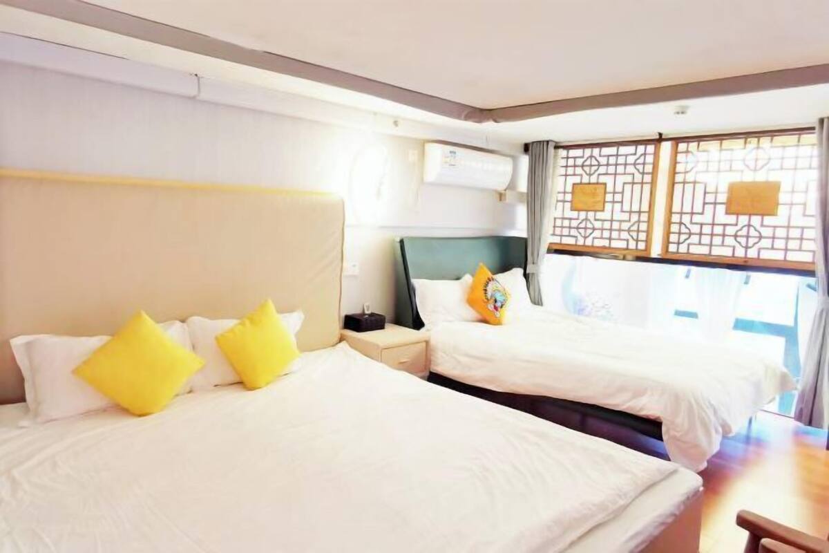 A hotel room with two beds, each with white bedding and yellow pillows, a nightstand with a lamp, and a window with a view.