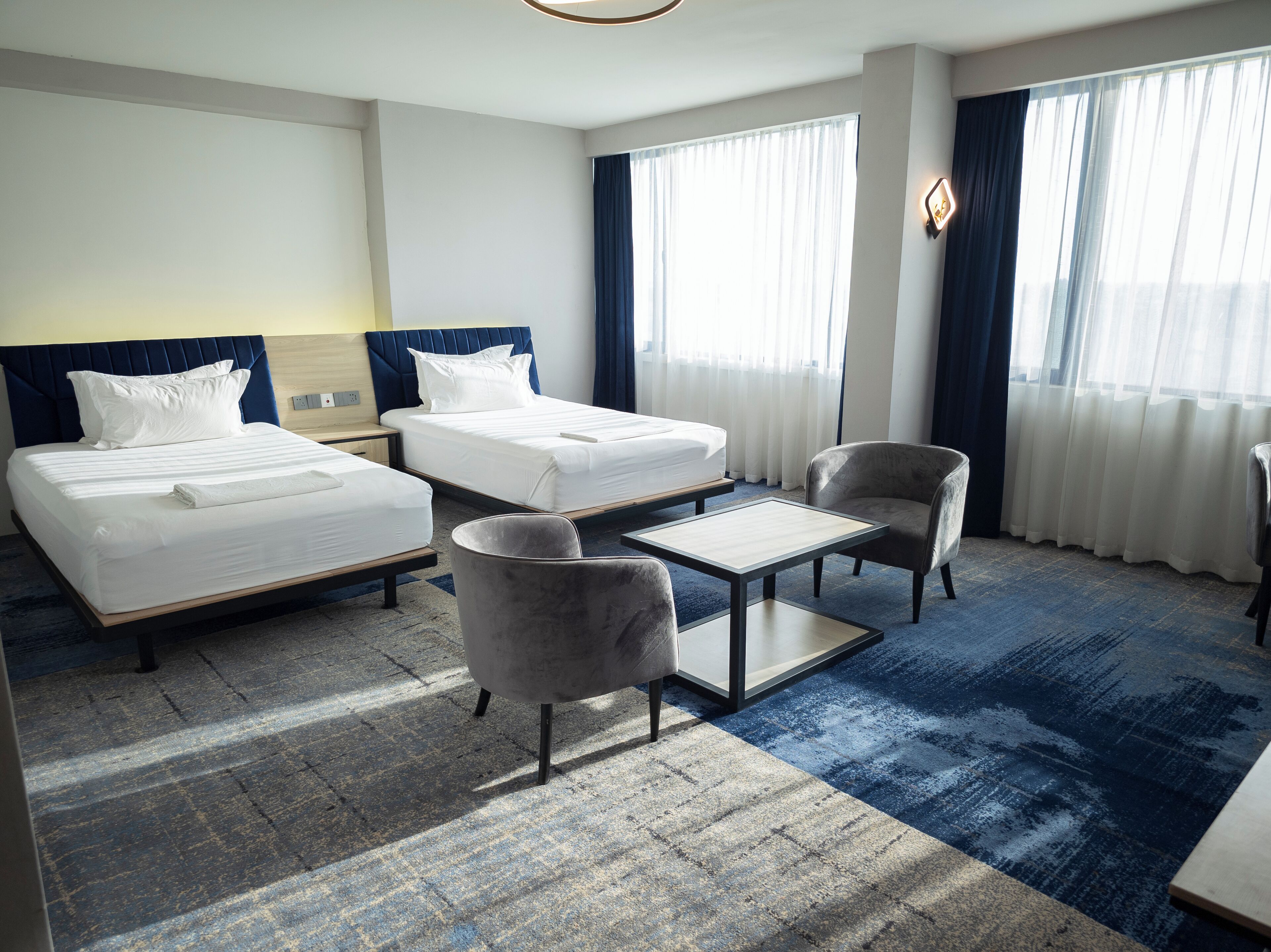 A hotel room with two beds, a small table, two armchairs, and a large window with curtains.