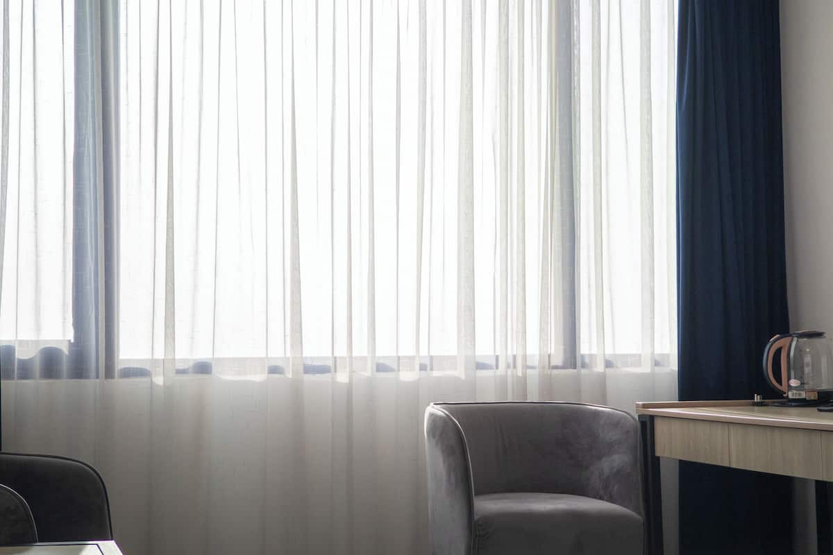 Una habitación con un ventanal cubierto por cortinas de gasa, un sillón gris, una mesita con una tetera y una vista parcial de una cama.