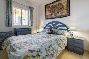 A bedroom with a bed, nightstands, a dresser, a window with curtains, and a wall hanging. - Apartment with communal pool (Mijas)