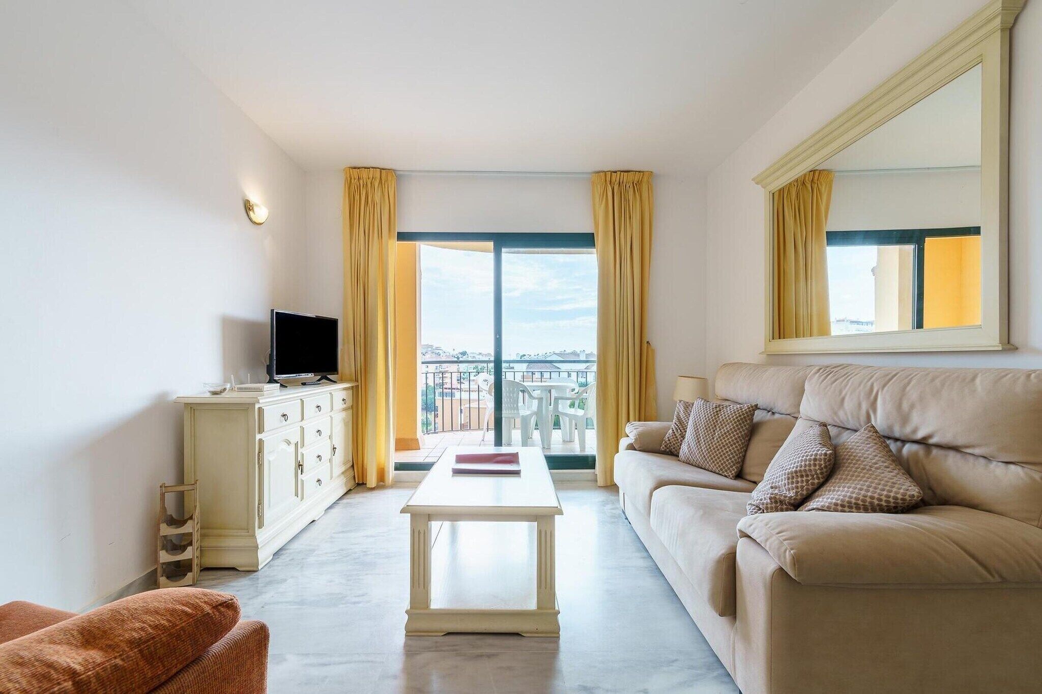 Ein Hotelzimmer mit einer Couch, einem Fernseher auf einer Kommode, einem kleinen Tisch und einem Balkon mit gelben Vorhängen.