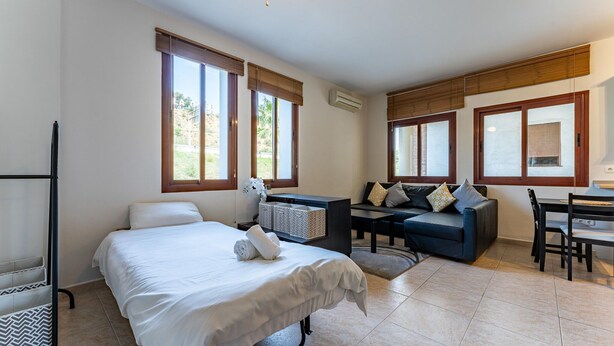 A hotel room with a bed, a sofa, a dining table, and a chair. There are large windows with blinds, an air conditioning unit, and a ceiling fan. - Apartment in preferred in Cala de Mijas (Mijas)
