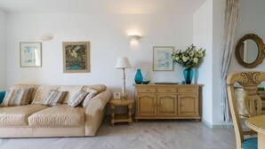 A living room with a beige sofa, a wooden cabinet, and a dining table with a chair. There are framed pictures on the wall and a vase with flowers on the cabinet. - Apartment in Mijas with pool (Mijas)