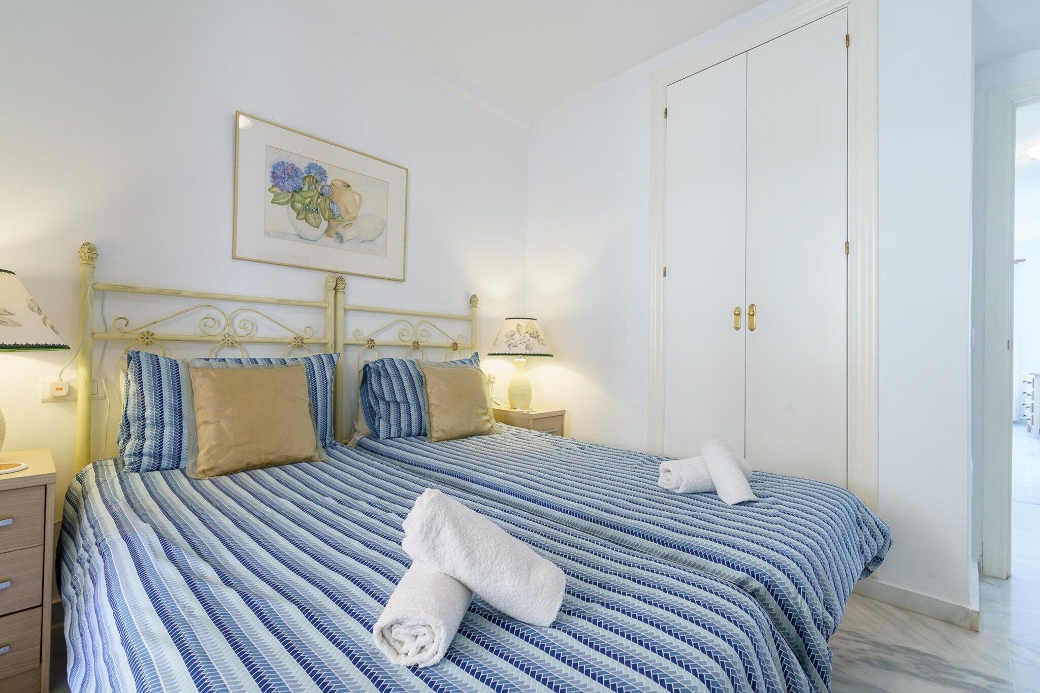 A neatly made bed with a striped blue and white bedspread, two pillows, and two rolled towels on the bed.
