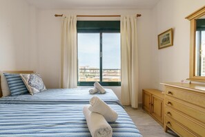 A bedroom with a bed, a dresser, a window with curtains, and a view of buildings outside.
