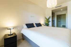 A modern bedroom with a large bed, a bedside table with a lamp, a potted plant, and a shower area visible through an open door. - Modern apartment in Mijas Costa (Mijas)