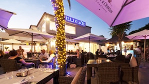 Un espace extérieur aménagé pour les repas, avec des meubles en osier et des parasols, une enseigne indiquant « OLIVIAE », et un bar en arrière-plan.
