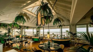 Une salle à manger spacieuse avec de hauts plafonds, de grandes fenêtres donnant sur l’océan et de nombreuses plantes suspendues.