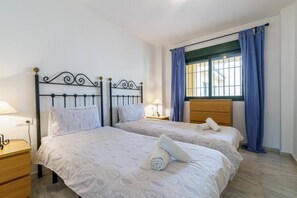 A hotel room with two beds, a wooden nightstand, a lamp, and a window with blue curtains.