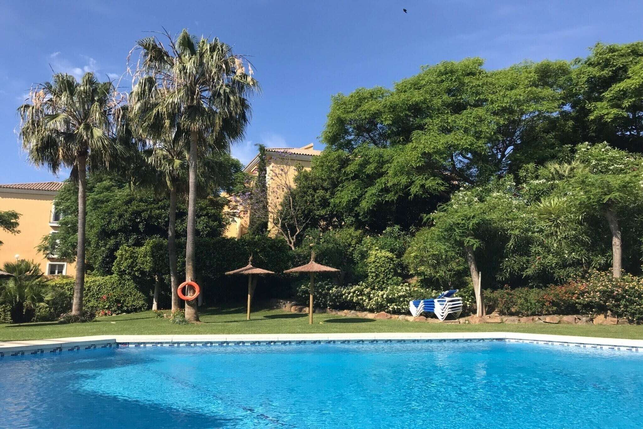 A swimming pool with palm trees and umbrellas, surrounded by lush greenery and a building in the background.