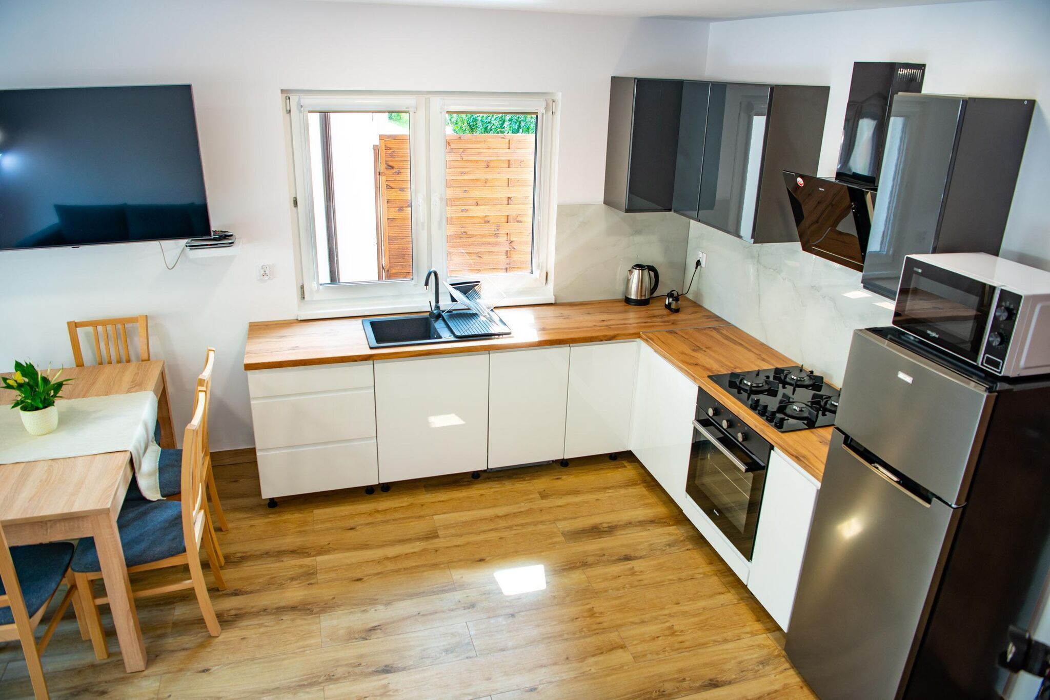 A modern kitchen with wooden flooring, white cabinets, a built-in oven, a microwave, and a sink.