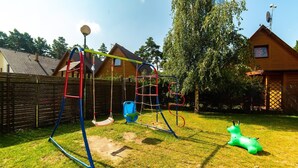 Un parque infantil con columpios y una estructura para escalar, rodeado por una cerca de madera y casas al fondo.
