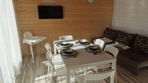 Interior de una cabaña de madera con una mesa de comedor preparada para cuatro personas, sillas de plástico blancas, un sofá con cojines estampados y un televisor montado en la pared.