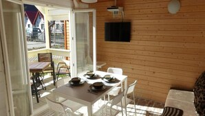 Un interior de madera con una mesa dispuesta para comer, sillas blancas y un televisor montado en la pared.