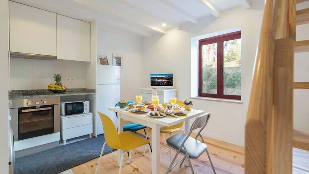 Et kompakt køkken med moderne hårde hvidevarer, et spiseområde med gule stole, og et vindue med udsigt til grøn bevoksning.