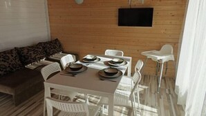 Una cabaña de madera con una mesa de comedor lista para cuatro personas, sillas de plástico blanco, un sofá con cojines estampados y un televisor montado.