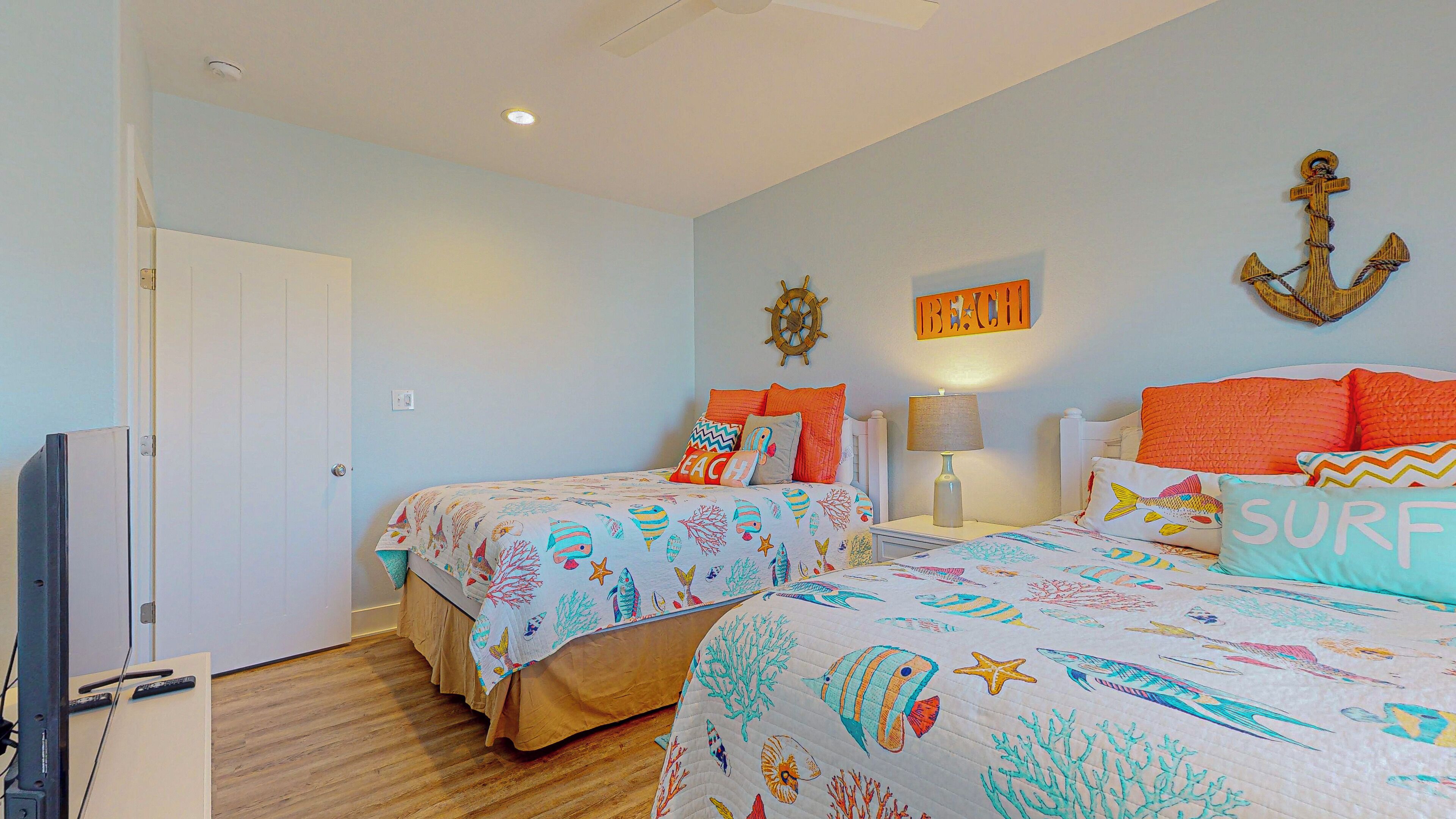 A bedroom with two beds, a wall-mounted anchor decoration, a 'BEACH' sign, and a 'SURF' pillow.
